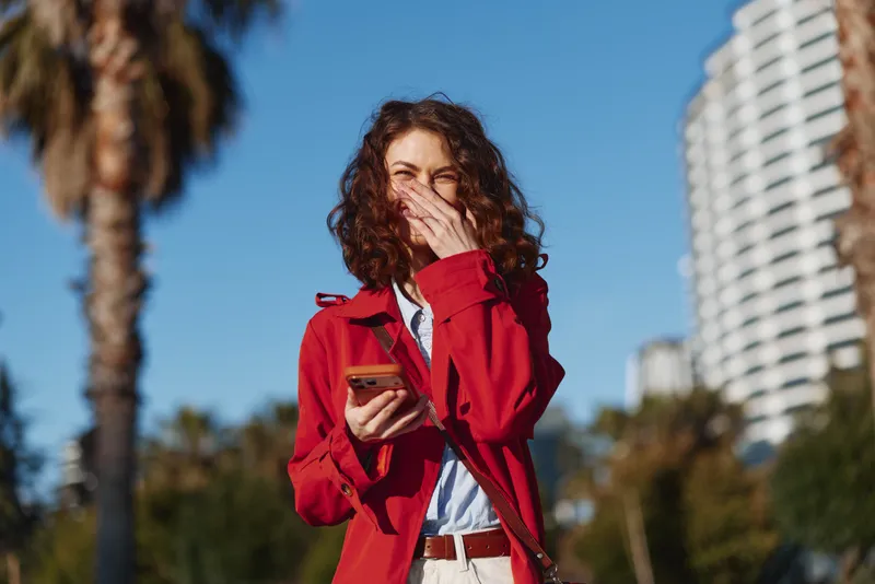 A surprised woman with curly hair in a red jacket checks her smartphone outdoors on a sunny day near palm trees and a tall modern building.