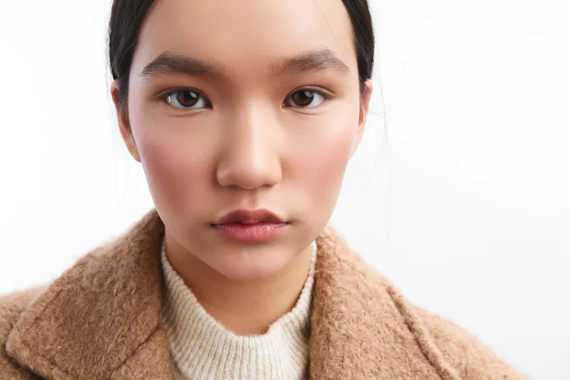 asian woman stares softly at the camera with a calm expression, wearing a neutral knit and tan coat, presenting a clean studio portrait with subtle lighting and tactile textures.
