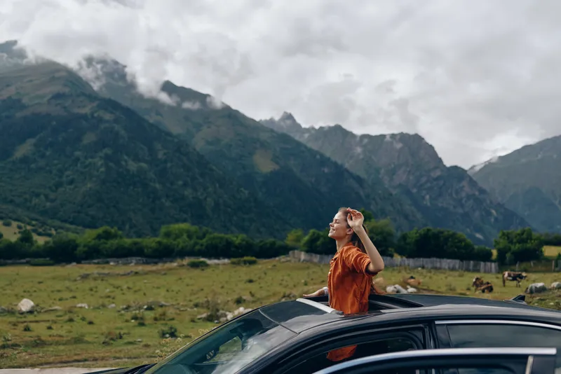 Woman stands on a car roof in a vast mountain landscape, wearing an orange shirt, admiring the view under cloudy skies while the open countryside stretches beyond.