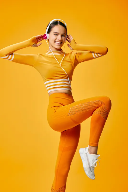 Woman in orange fitness outfit dances mid-air with headphones against a bright orange background, showcasing dynamic movement, athletic style and energetic sport lifestyle.