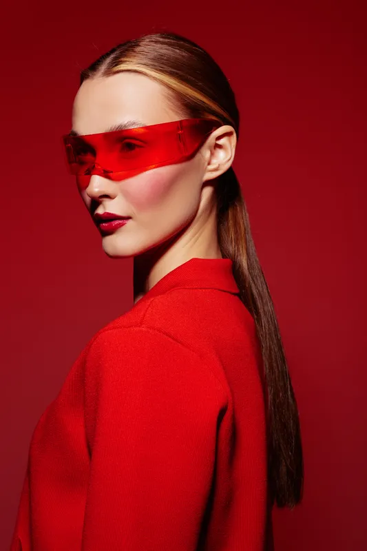 Stylish woman with long hair wearing red futuristic sunglasses and red textured jacket poses on red background. Concept for fashion, modern style, and bold color trends.