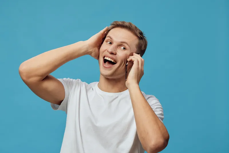 Young Caucasian man wearing a casual white t-shirt, laughing and holding his hands near his ears, looking joyful and cheerful, isolated on a plain turquoise background. People lifestyle concept, happi