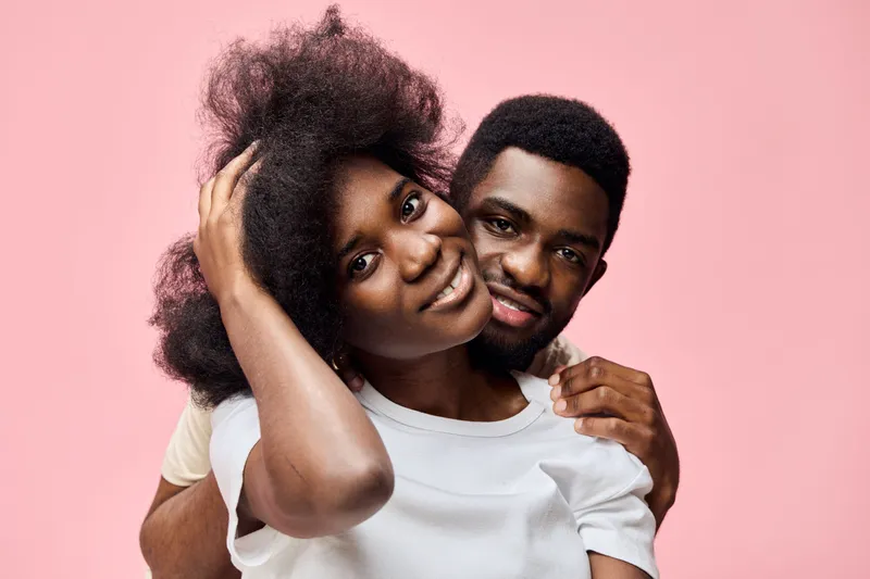 Happy young African couple smiling together with playful poses against a soft pink background, representing love and joy in relationships
