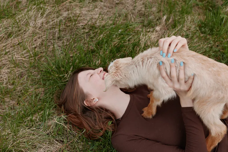 A woman lies on grass outdoors, pets a soft goat kid with gentle affection, smiles as the animal nuzzles her, capturing a warm lifestyle moment of connection and relaxation in a natural setting