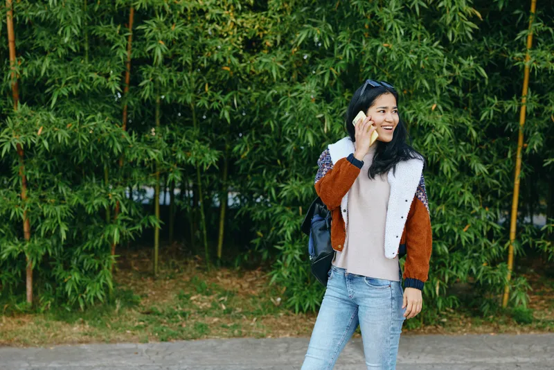Woman talking on phone while walking outdoors along a street, wearing a color block jacket and jeans, smiling with trees in the background, fashion lifestyle moment