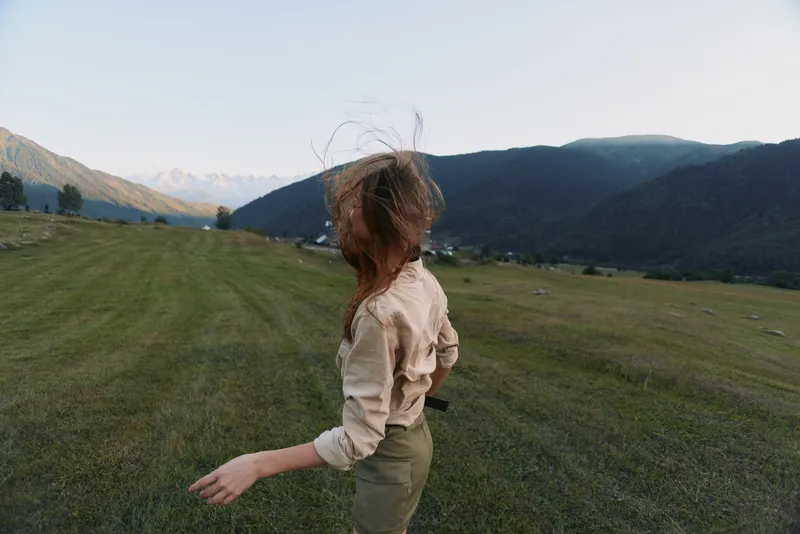 Woman in casual clothing on a grassy hillside with distant mountains, wind in her hair, bright sky. Outdoor lifestyle, nature, and natural beauty.
