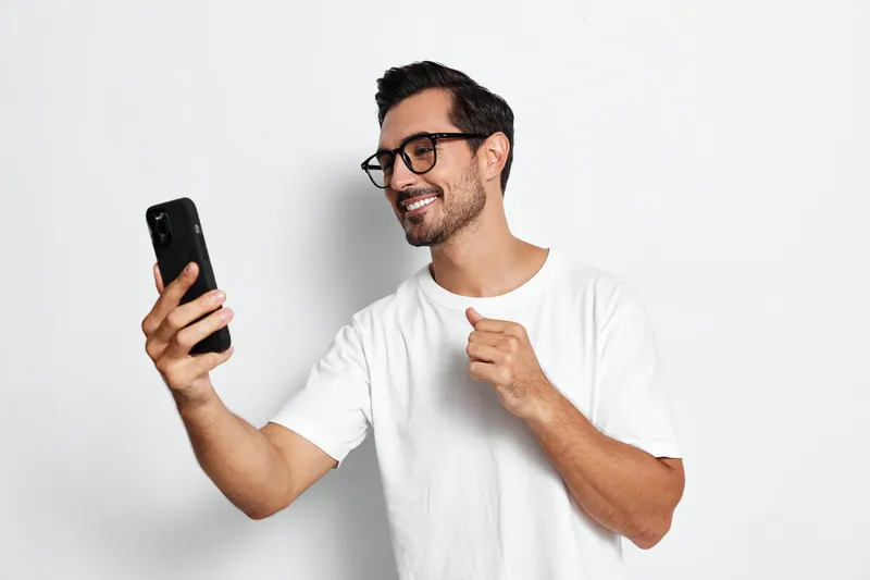 Smiling young man wearing glasses holding a smartphone and looking at the camera with excitement, isolated on plain white background, joyful lifestyle concept.