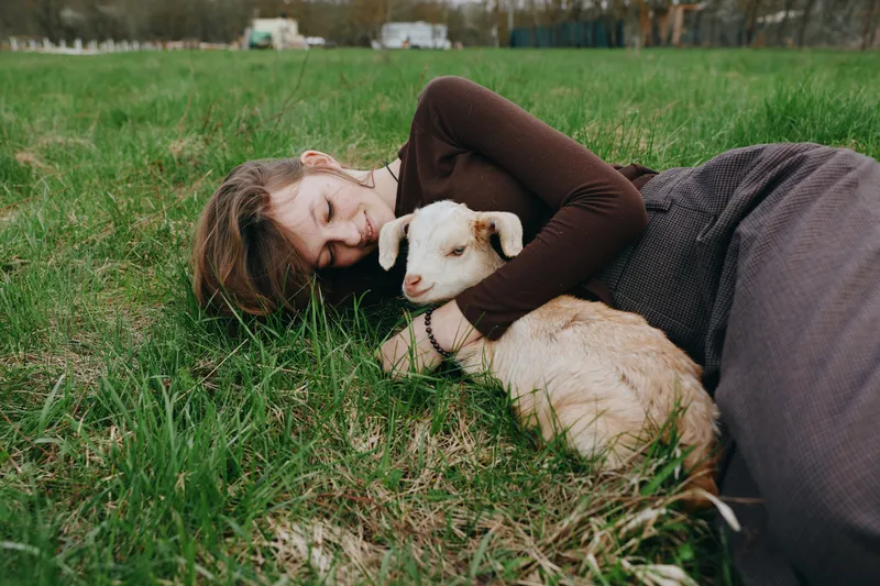 Joyful woman outdoors, lying on grass, cuddling a goat kid, sunlit moment, relaxed posture, gentle breeze, natural colors, pastoral setting, candid lifestyle in a peaceful countryside field.