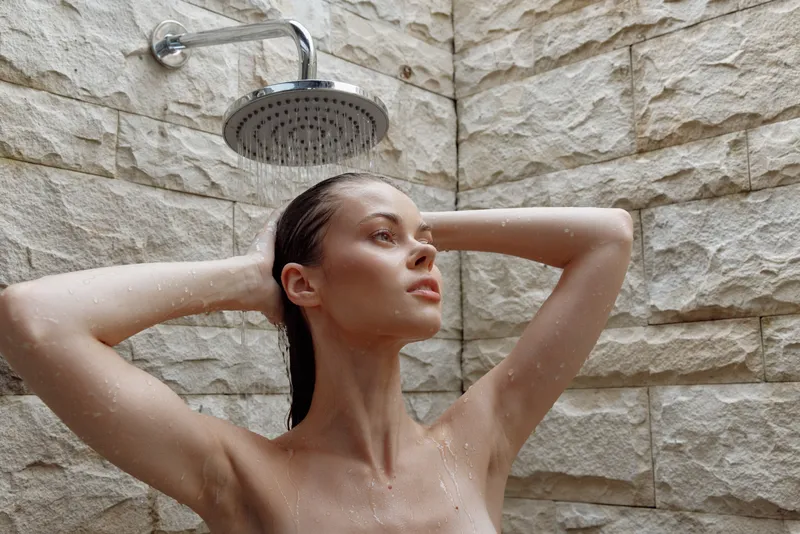 young woman taking a shower indoors with water droplets, relaxed expression, wet hair, beige textured stone wall background