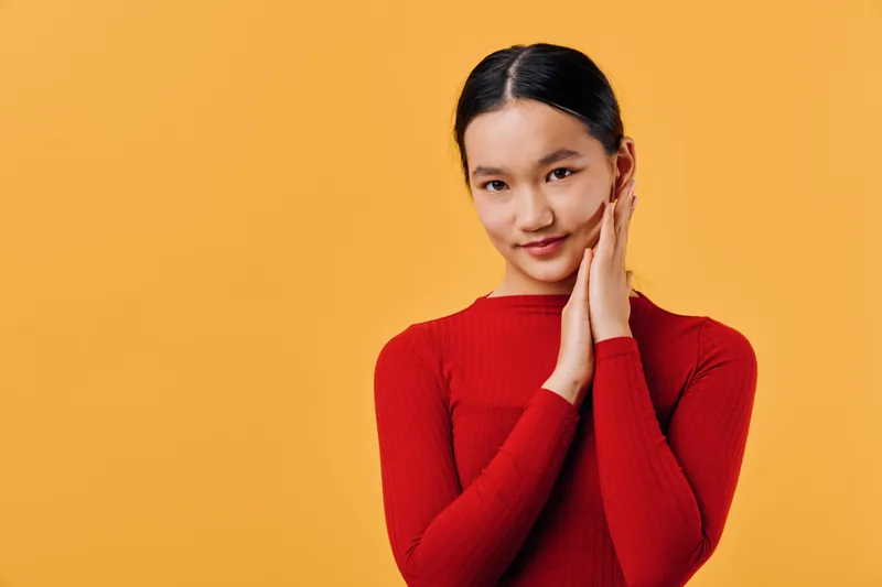 Young woman with dark hair and red sweater posing with hands together near face smiling confidently on orange background studio portrait of female model with clear skin and neat hairstyle.