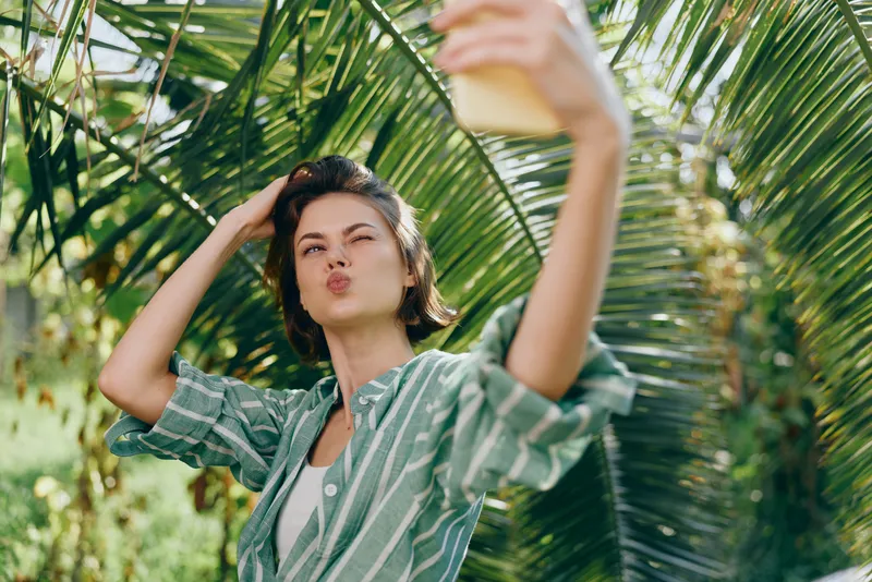 Young woman takes a selfie in a tropical setting, wearing a striped green shirt, puckering lips for the phone photo.