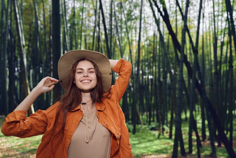 Woman in straw hat with a bright smile in bamboo forest nature portrait, happy young female wearing orange jacket enjoying outdoors, sunlight, casual bohemian lifestyle and relaxed mood.