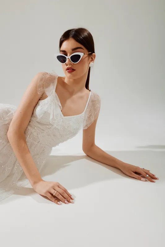 Young woman in a white lace dress wearing sunglasses poses lying on a white surface. The image focuses on beauty, fashion, and modern style with soft lighting.