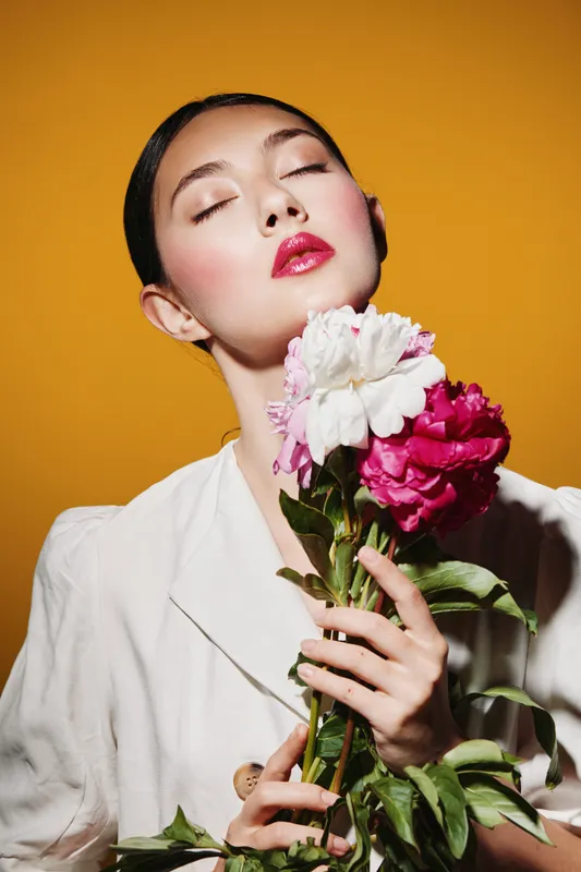 Young woman with glossy pink lips and flawless skin holds bright flowers close while her eyes gently close against a vibrant yellow backdrop. Beauty, fashion, creative studio portrait.