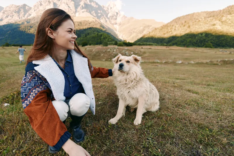 Woman petting dog in mountains meadow, smiling and crouching beside her pet in outdoor countryside scene with autumn light. Happy owner in warm jacket with furry companion.