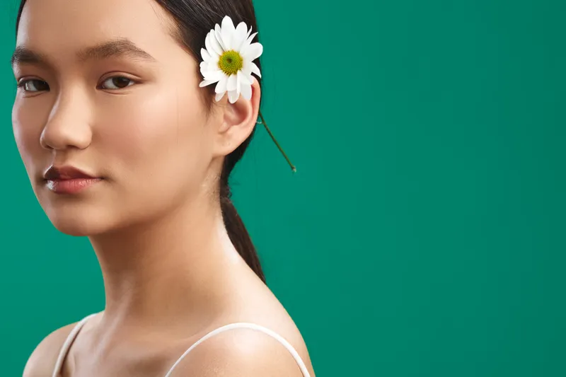 Asian woman with a white flower in her hair stands against a vivid green backdrop, exuding calm poise and confidence, inviting focus, creativity, and positive energy in the scene.