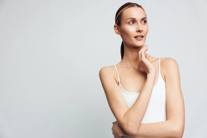 Young woman with fair skin and pulled back hair wearing white tank top posing thoughtfully against light gray background with soft natural lighting and minimalistic style