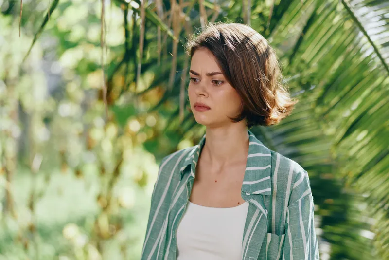 A young woman stands in a sunny garden among tropical leaves, wearing a green striped jacket over a white top.