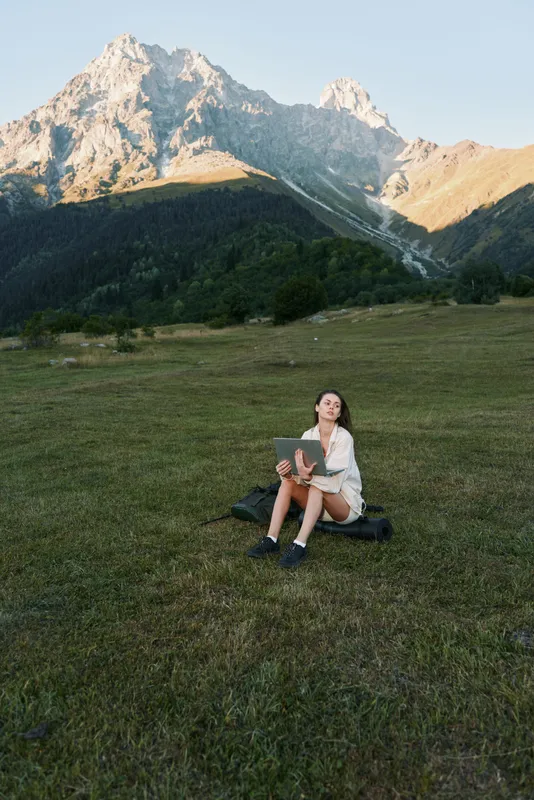 A woman sits on a grassy meadow with a laptop in a rugged mountain landscape. She wears casual clothes and looks focused, enjoying productive remote work in a serene outdoor setting.