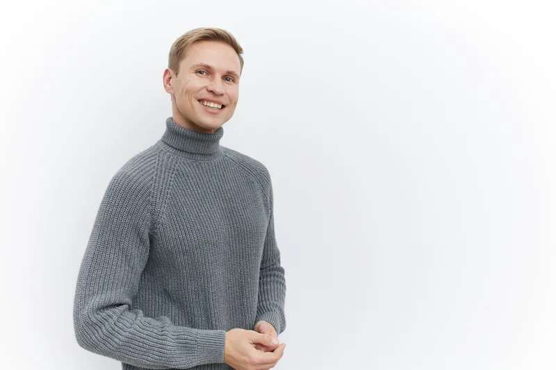 Smiling young man wearing a cozy gray turtleneck sweater looks happily at the camera in a studio with plain white background, creating a friendly and approachable atmosphere, suitable for lifestyle an