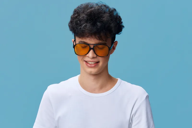 young man wearing orange sunglasses and white t-shirt smiling against blue background in casual summer style with curly hair and clear skin