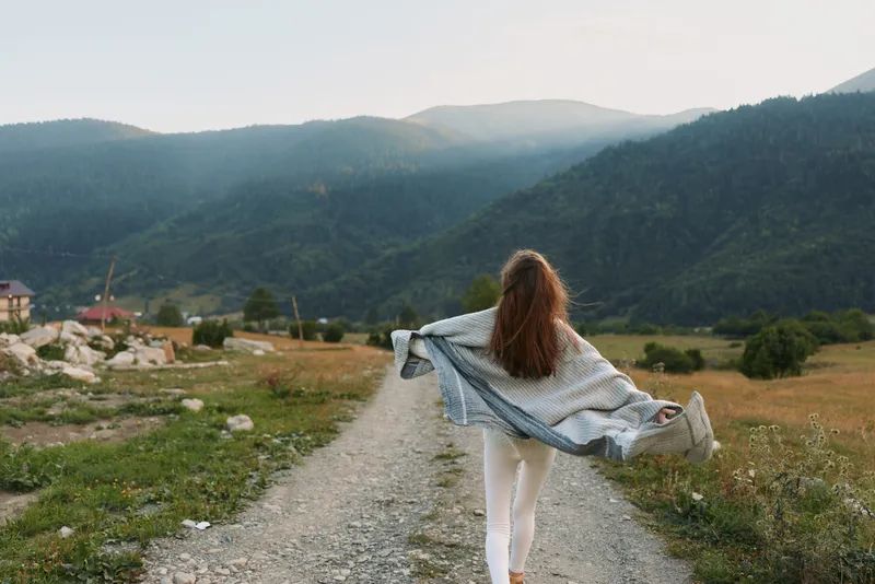 Cheerful woman outdoors, walking along a rustic path in a mountainous landscape, scarf fluttering, sunlit breeze, casual lifestyle, natural colors, peaceful countryside backdrop amid mountains.