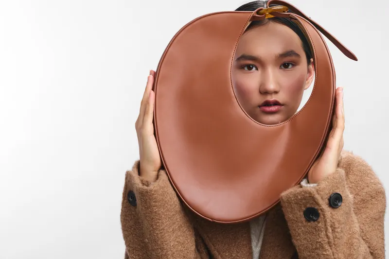 asian woman wearing a warm coat holds a circular accessory to her face, creating a stylish studio portrait with a clean backdrop and a subtle, engaging gaze.