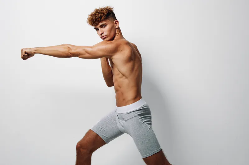Young man with curly hair in gray shorts practicing boxing punch against white background. Athletic shirtless male training for fitness and strength with focused expression.