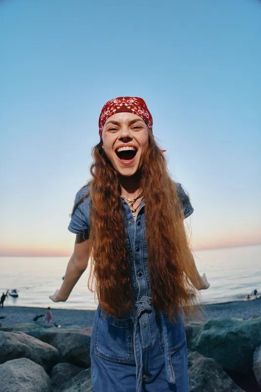 lifestyle woman in modern boho-western denim street style with long hair and red bandana smiling joyfully at seaside, film color effect enhancing vibrant mood
