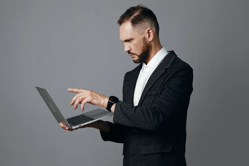 A focused professional man in business attire uses a laptop against an isolated gray background, conveying confidence and concentration as he analyzes data and communicates ideas with precision.
