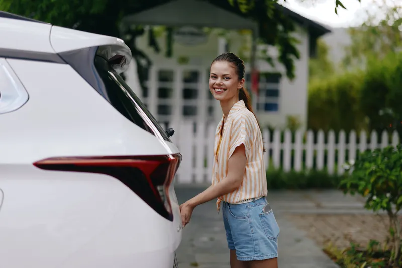Улыбающаяся молодая женщина стоит рядом с белым автомобилем, создавая живую и позитивную атмосферу