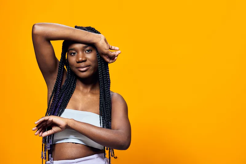 A young Black woman with braided hair poses confidently against a vibrant yellow backdrop, showcasing her stylish outfit and expressing joy and empowerment