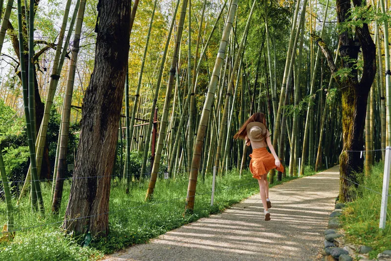 Woman running along a gravel path through a dense bamboo forest wearing a sun hat and orange skirt, nature trail with sunlight on green trees, motion and carefree summer travel mood.