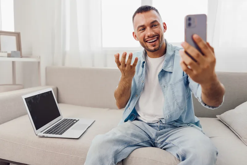 Happy man engaging in a video call, sitting comfortably on a couch, wearing casual attire, showcasing a bright and modern interior with a laptop nearby.