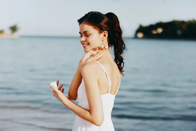 Young woman in a white sundress stands at the shoreline, turning with a gentle smile as sea breeze lifts her hair, embodying calm coastal beauty and carefree summer bliss.