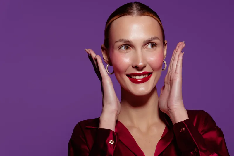 Smiling woman with red lipstick and earrings poses with hands near face on purple background. Concept of beauty, makeup, fashion, and confident female model in studio lighting.