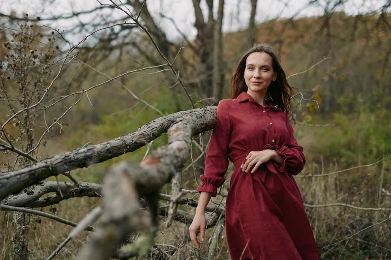 A genuine portrait of a woman in a simple red dress leaning on a fallen tree in an overgrown field, capturing natural expression, authentic presence and honest storytelling.
