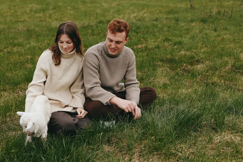 Young couple wearing knitted sweaters sits on green grass in a field while a white goat kid explores nearby, creating a relaxed and natural lifestyle scene in the countryside.