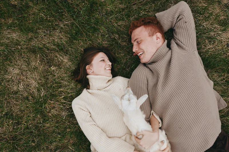 Happy couple lying on grass in cozy sweaters, smiling at each other, a small dog between them, outdoor lifestyle moment on a sunny day, intimate candid scene of affection