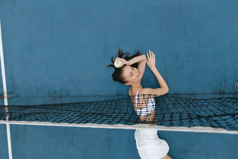 Joyful young Asian woman relaxing on a tennis court, showcasing sporty fashion in a vibrant blue setting with a playful attitude and fresh summer vibes