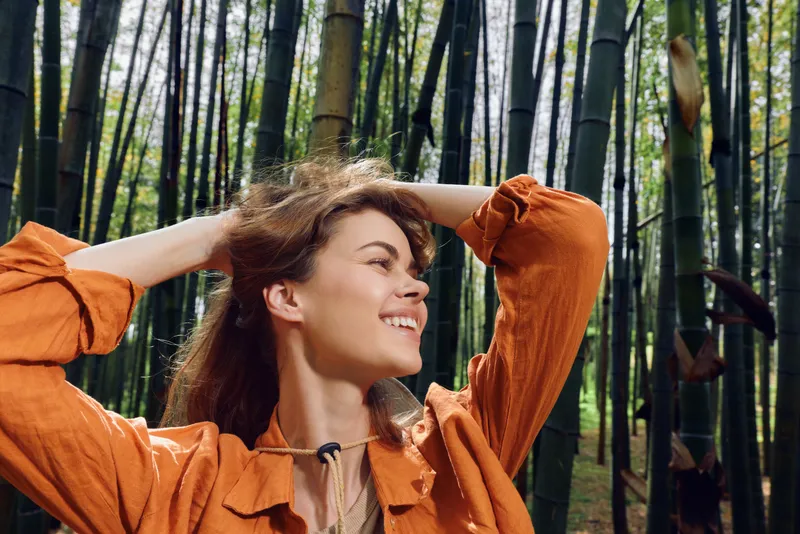 Woman in bamboo forest smiles and looks upward while adjusting hair, outdoor portrait of a happy young female in nature wearing orange jacket, relaxed and joyful scene.
