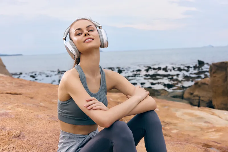 Young woman enjoying music with headphones at the seaside, showcasing a relaxed and joyful expression, wearing sporty activewear against a tranquil ocean backdrop