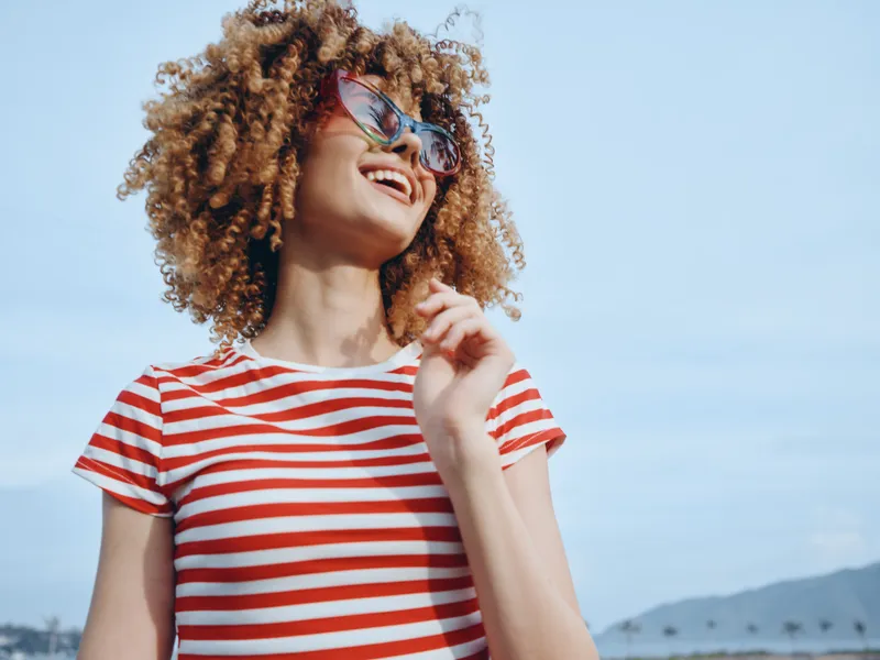 Smiling woman with curly hair wearing a red and white striped shirt and sunglasses, standing outdoors under a clear blue sky. Her joyful expression captures a bright, carefree summertime moment.