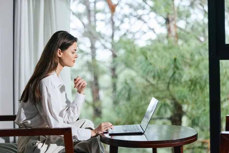 Задумчивая женщина в удобной одежде занимается удаленной работой за ноутбуком на фоне безмятежной природы, создающей умиротворяющую атмосферу для продуктивной деятельности