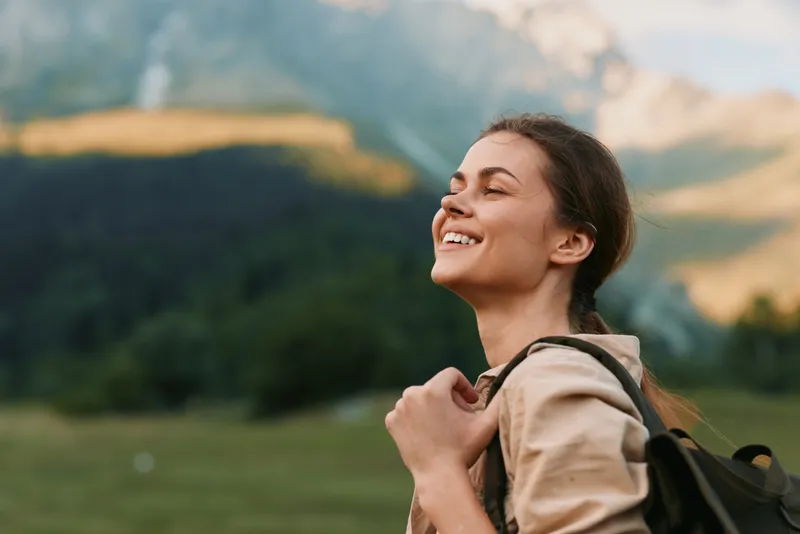 Smiling woman with backpack enjoying fresh air outdoors in nature. Relaxed lifestyle concept with mountains and green landscape in the background. Bright daylight.