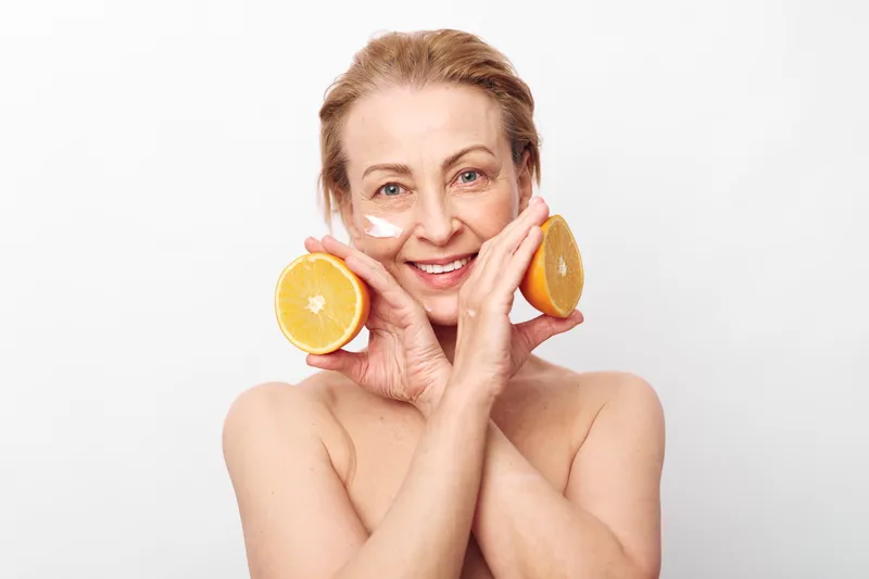 Smiling middle aged woman with natural skin holds two halves of fresh orange near face with cream applied under eyes. Concept for skincare, healthy lifestyle, natural beauty, and wellness.