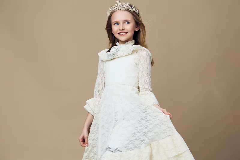 Elegant girl wearing a delicate white lace dress and tiara, smiling against a soft beige background, embodying childhood joy and innocence in a timeless style