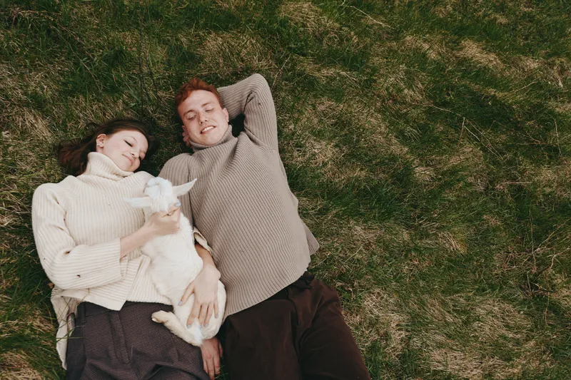 Couple lying on grass outdoors, relaxed smiles, cuddling a playful goat kid, cozy sweaters, sunlit calm, natural colors, gentle breeze, warm bond, candid moment, enjoying a peaceful afternoon