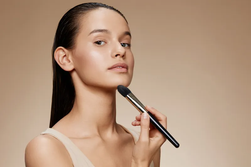 Young woman applying makeup with a brush, showcasing natural beauty and skincare routine, perfect for beauty and cosmetic promotions