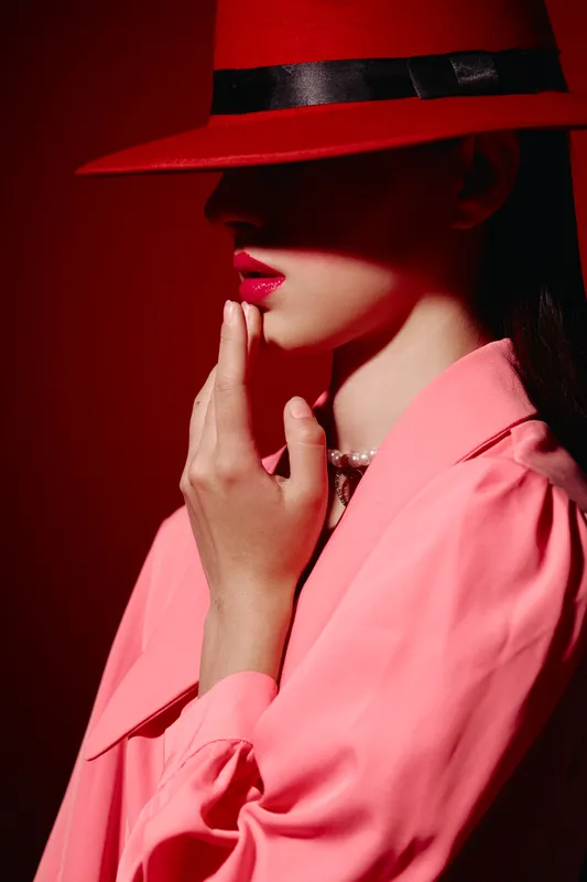 Fashion woman in a red hat and pink coat poses with softly lit face almost hidden in shadow against a red background. Concept for beauty creative and stylish portrait.